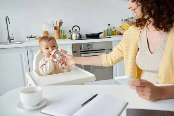 Frau mit Handy hilft kleinem Mädchen beim Trinken aus Babyflasche während der Arbeit in der Küche — Stockfoto