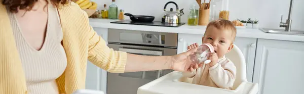 Caring mother helping toddler girl drinking from baby bottle in kitchen, banner — Stock Photo