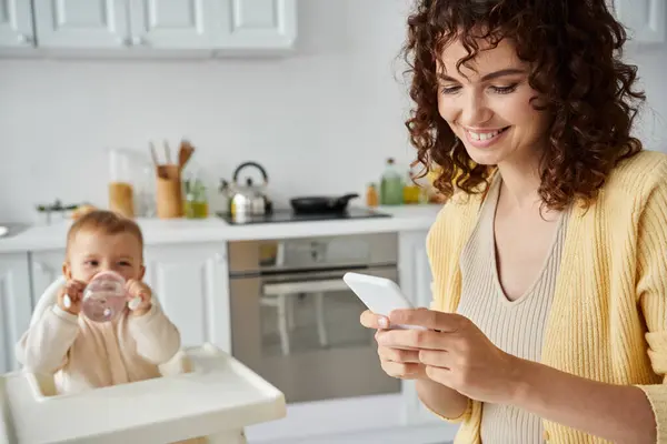 Mensajería mujer sonriente en el teléfono móvil, mientras que el niño bebiendo de biberón en la cocina - foto de stock