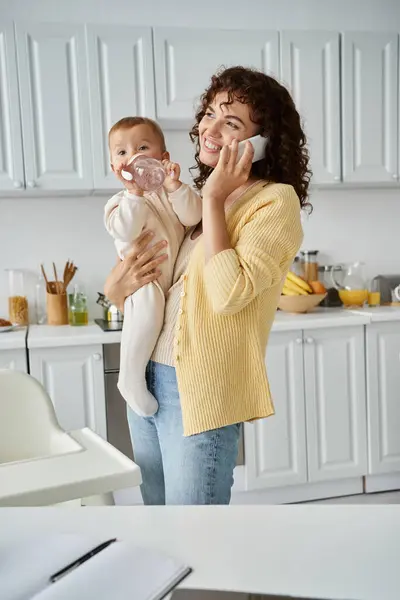 Fröhliche Mutter spricht mit Smartphone und hält Kind trinkend aus Babyflasche in Küche — Stockfoto