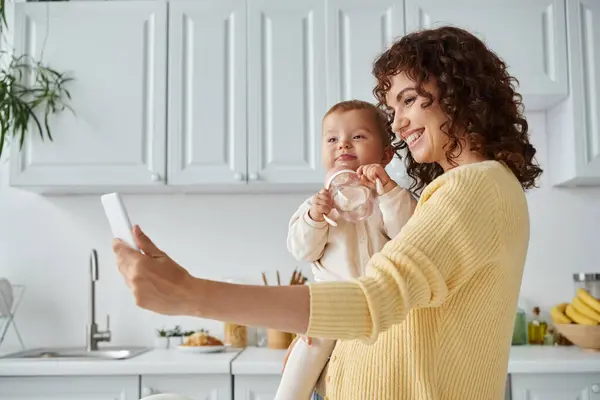 Madre emocionada tomando selfie en el teléfono inteligente con niño pequeño sosteniendo biberón, momento sincero - foto de stock
