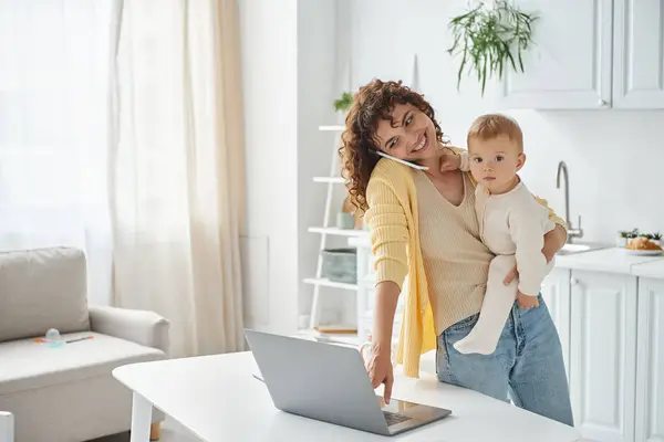 Busy mother talking on smartphone and using laptop while holding toddler daughter in kitchen — Stock Photo
