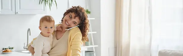 Trabalho freelance, mulher sorridente segurando criança enquanto conversa no smartphone na cozinha, banner — Fotografia de Stock