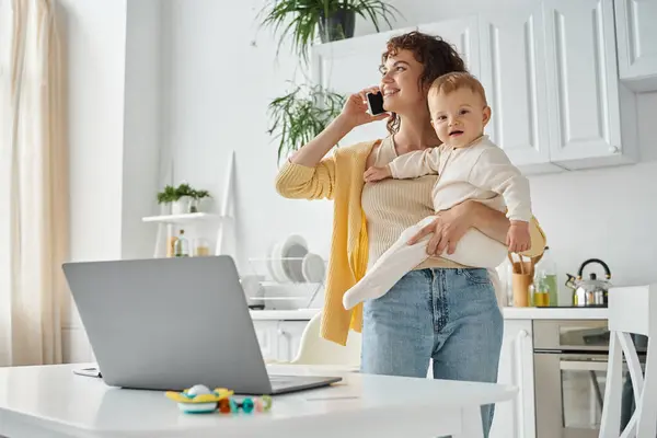 Career and parenthood, happy woman talking on smartphone and holding baby near laptop in kitchen — Stock Photo