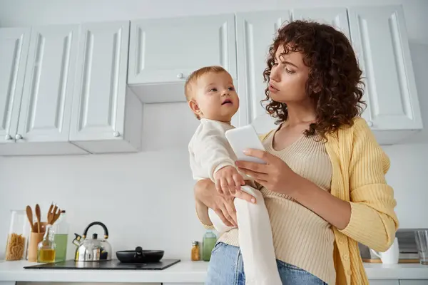 Кучерява жінка тримає дочку малюка і мобільний телефон в руках, стоячи на сучасній кухні — стокове фото