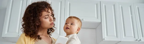 Woman with little daughter and mobile phone in hands standing in modern kitchen, modern life, banner — Stock Photo