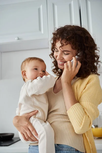 Donna felice che tiene adorabile bambina e parla sullo smartphone in cucina, beata maternità — Foto stock