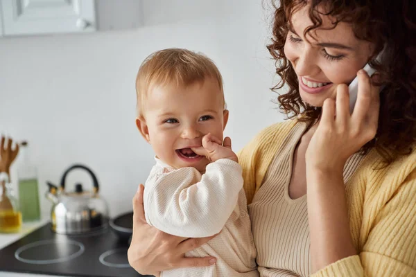 Sonriente mujer sosteniendo alegre niño hija y hablando en el teléfono inteligente en la cocina, felicidad - foto de stock