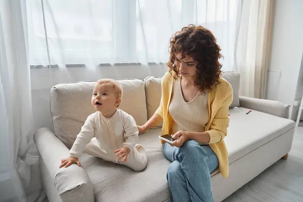 Woman networking on mobile phone near toddler daughter on cozy couch in modern living room, leisure — Stock Photo