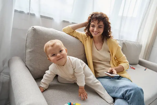 Glückliche Mutter sitzt mit Smartphone neben entzückter kleiner Tochter auf Couch im Wohnzimmer — Stockfoto