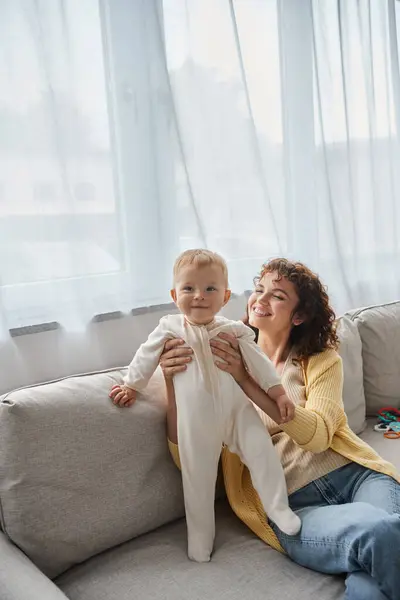 Mujer llena de alegría sosteniendo niña sonriente en el sofá en la acogedora sala de estar, tiempo de calidad en casa - foto de stock