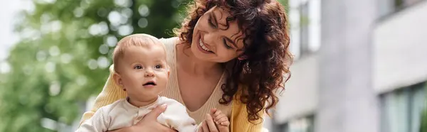 Femme aux cheveux ondulés souriant près de mignon tout-petit fille dans la rue de la ville, maternité heureuse, bannière — Photo de stock