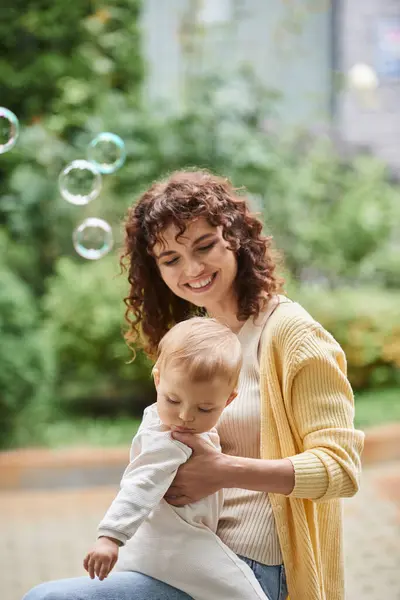 Porträt einer fröhlichen Frau mit kleiner Tochter in der Nähe fliegender Seifenblasen — Stockfoto