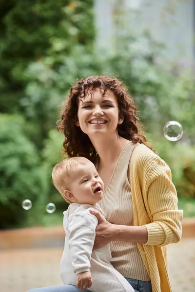 Retrato de mujer excitada con bebé hija cerca de burbujas de jabón volador al aire libre, momentos felices - foto de stock