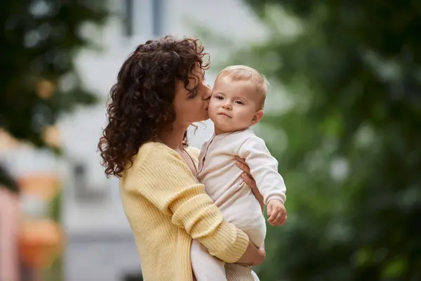 Femme aux cheveux ondulés embrassant et embrassant mignonne petite fille à l'extérieur, maternité heureuse — Photo de stock