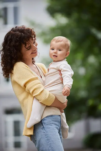 Freudige Frau mit welligem Haar, die entzückendes Baby hält, während sie draußen steht, Qualitätszeit — Stockfoto