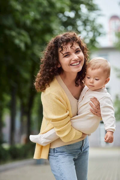 Curly woman smiling at camera while holding little daughter on urban street, happy modern family — Stock Photo