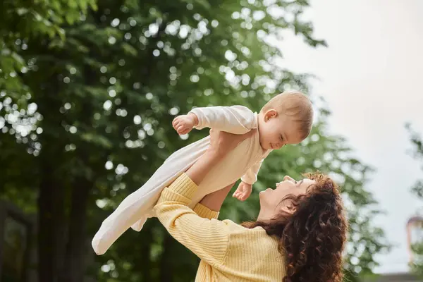 Mujer emocionada criando a su hijita en las manos mientras se divierte al aire libre, maternidad feliz - foto de stock
