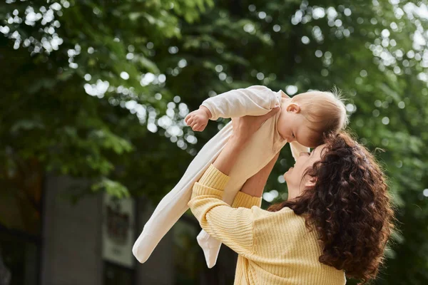 Feliz madre jugando con un niño pequeño mientras la crían en las manos al aire libre, el ocio y la emoción - foto de stock