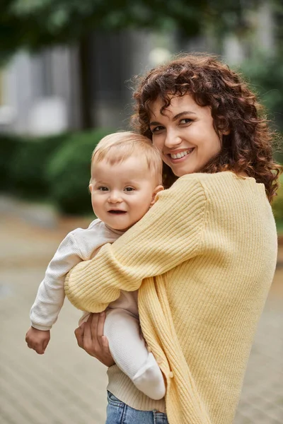 Overjoyed woman with wavy hair embracing toddler daughter and smiling at camera outdoors, happiness — Stock Photo