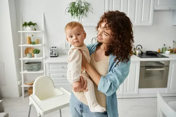 Donna allegra che tiene adorabile figlioletta in pagliaccetto mentre in piedi in cucina moderna — Foto stock