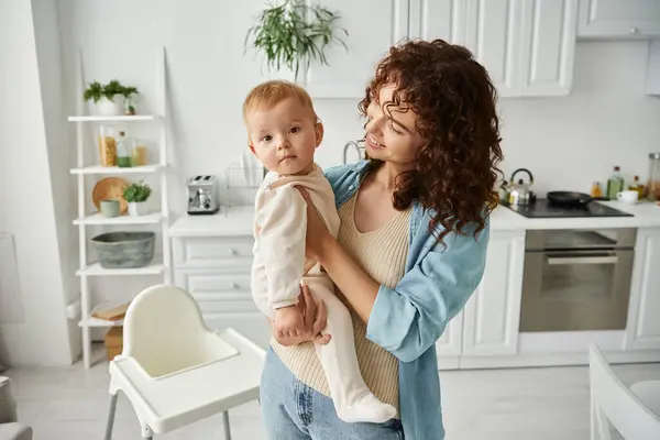 Cheerful woman holding adorable little daughter in romper while standing in modern kitchen — Stock Photo