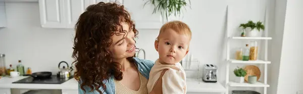 Woman with wavy hair smiling near cute baby girl in kitchen, happy motherhood, horizontal banner — Stock Photo