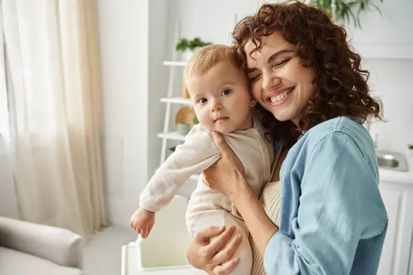 Mujer llena de alegría con los ojos cerrados sosteniendo y abrazando adorable niña en casa, la paternidad feliz - foto de stock