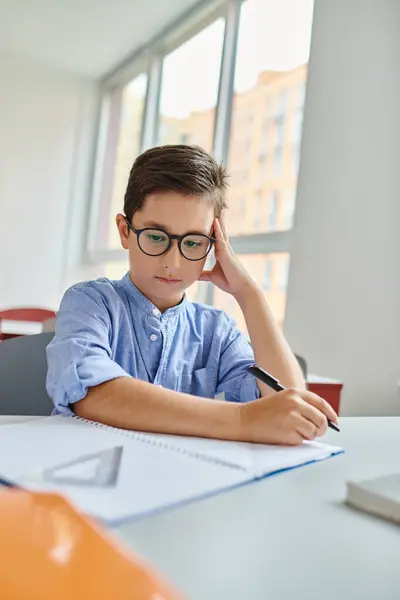 A young boy with a pen in hand, focused and engaged, sitting at a desk with paper in a vibrant classroom setting. — Stock Photo