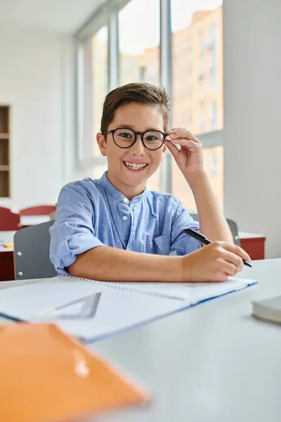 A young boy with glasses sits attentively at a table in a colorful classroom — Stock Photo