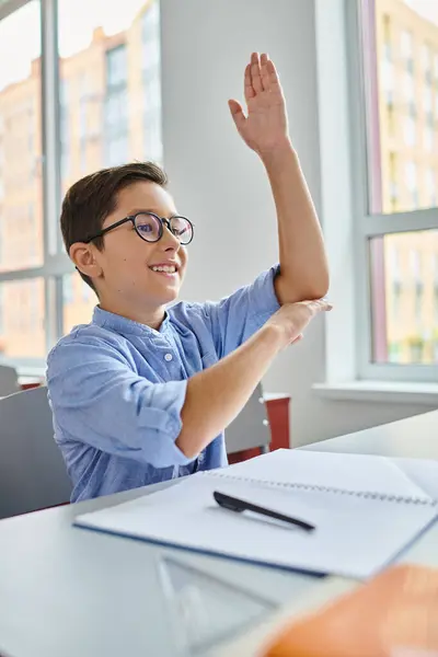Ein kleiner Junge sitzt an einem Schreibtisch in einem hellen, lebhaften Klassenzimmer und hebt die Hand, um am Unterricht teilzunehmen.. — Stockfoto