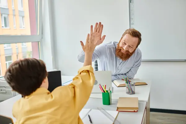 Un niño con una camisa amarilla le está dando cinco a un profesor en un ambiente brillante y animado en el aula. - foto de stock