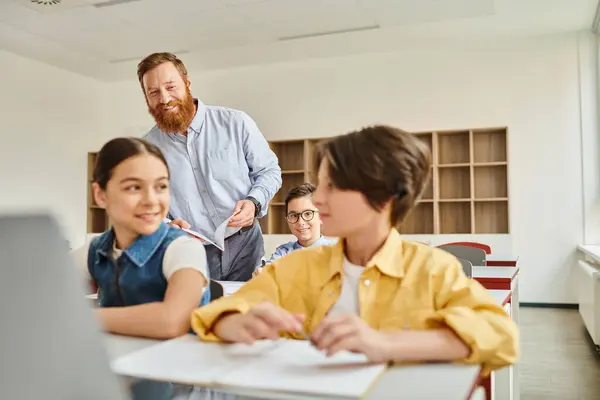 Un grupo de niños se sientan atentamente en los escritorios de una clase iluminada, absortos en la lección de computación que les enseña su maestro masculino. - foto de stock