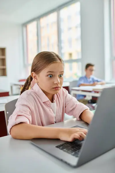 Una giovane ragazza con i capelli intrecciati si siede a una scrivania, focalizzata su un computer portatile in una vibrante aula — Foto stock