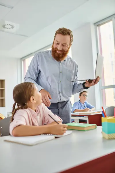 Un homme se tient à côté d'une petite fille dans une salle de classe animée, s'engageant dans une discussion individuelle pendant que le reste du groupe d'enfants apprend activement autour d'eux. — Photo de stock