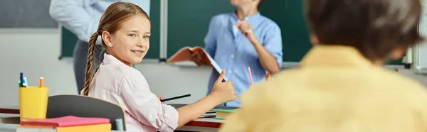 Une jeune fille s'assoit avec attention à un bureau devant un tableau noir, écoutant les instructions de ses professeurs dans un cadre de classe lumineux. — Photo de stock