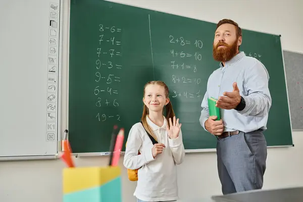 Un uomo si trova accanto a una bambina davanti a una lavagna, insegnandola e guidandola in un ambiente di classe luminoso e vivace. — Foto stock