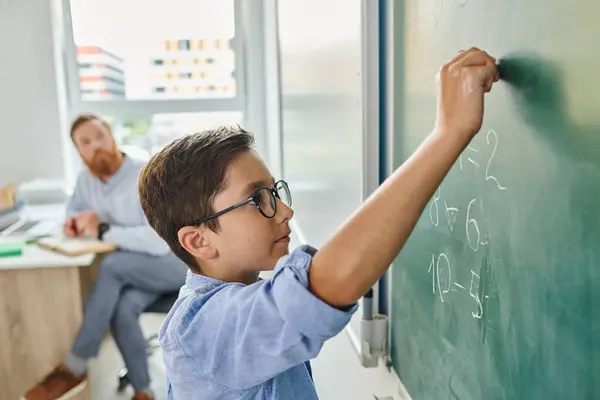 A young boy engrossed in writing on a blackboard while a man looks on in a bright, lively classroom setting. — Stock Photo