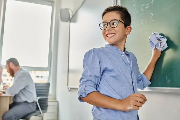 Kid stands before a blackboard, smiling in a vibrant classroom setting. — Stock Photo
