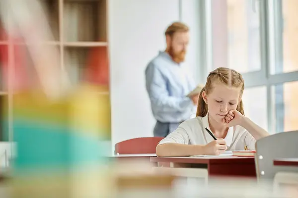Une jeune fille est assise à un bureau, engagée dans une étude dans une salle de classe lumineuse et animée pendant que l'enseignant enseigne derrière elle — Photo de stock