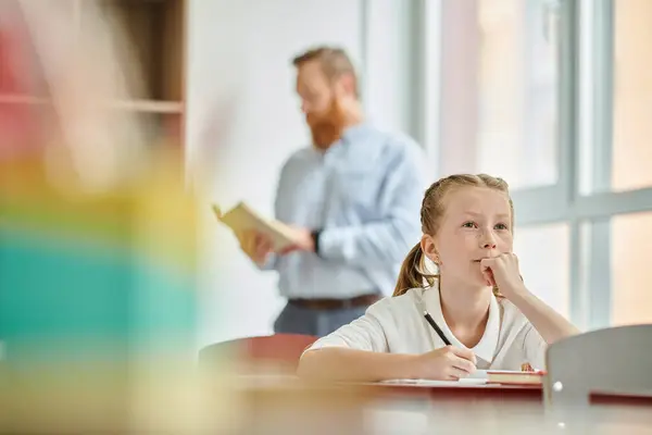 Молодая девушка сидит за столом, в то время как мужчина учитель стоит позади нее, инструктируя класс — стоковое фото