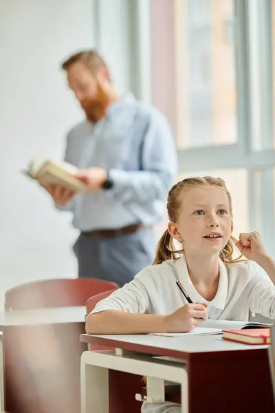 Une petite fille s'assoit attentivement à son bureau, absorbant une leçon d'un homme dans un cadre de classe dynamique et accueillant. — Photo de stock