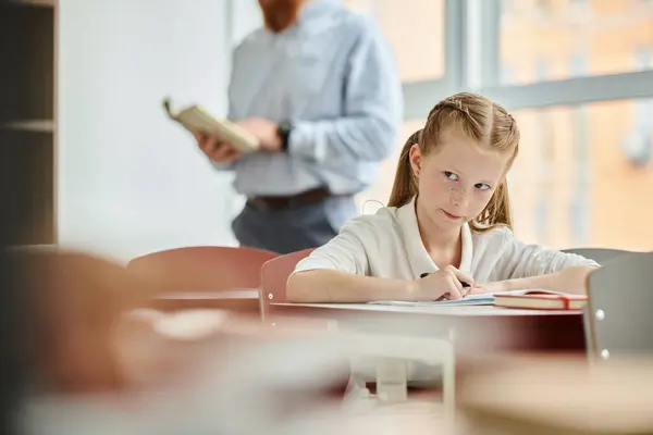 Молодая девушка, обнимающая школьный мир, сидя за столом в ярком классе. — стоковое фото