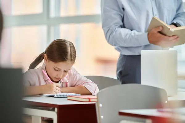 Una giovane ragazza si siede alla scrivania, avvolta in un libro e utilizzando una calcolatrice. L'aula è luminosa e vivace, con un insegnante di uomo che la istruisce — Foto stock