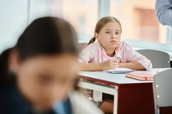 Ein junges Mädchen mit Zöpfen sitzt an ihrem Schreibtisch und hört den Anweisungen der Lehrer in einem geschäftigen Klassenzimmer zu.. — Stockfoto