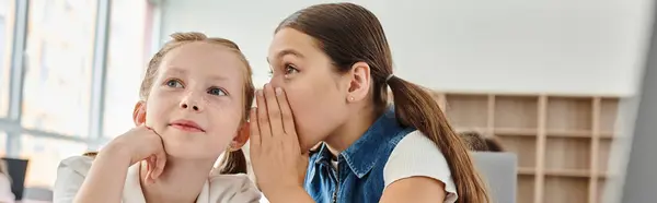 Due bambine sedute a un tavolo, che spettegolano in un'aula vivace. — Foto stock