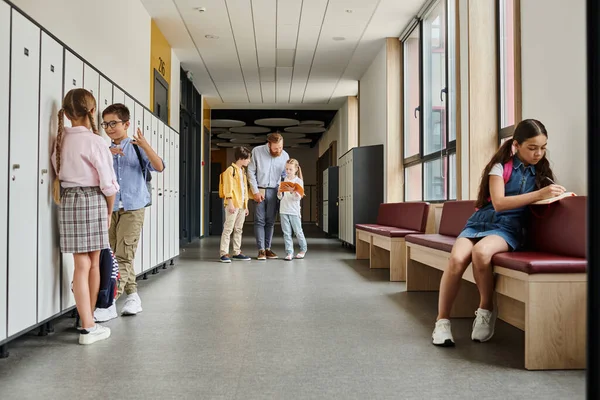Groupe d'enfants debout dans le couloir à côté des casiers tandis qu'un enseignant les instruit dans un cadre de classe lumineux et animé. — Photo de stock