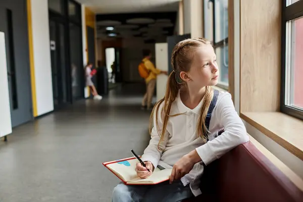 A young girl with a focused expression sits on a window sill, writing in a notebook as sunlight streams in through the glass pane. — Stock Photo