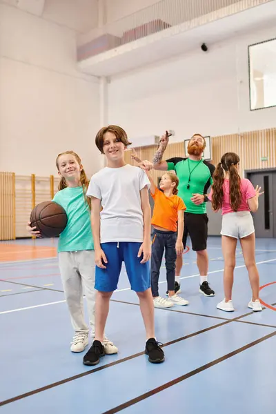 Un gruppo eterogeneo di bambini piccoli si trova fiduciosi in cima a un campo da basket, immersi nell'energia del momento mentre ascoltano attentamente le istruzioni dei loro insegnanti maschi.. — Foto stock