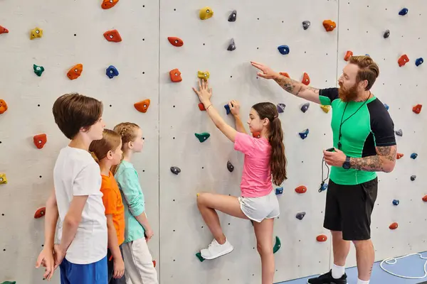 Un insegnante di sesso maschile assiste un gruppo eterogeneo di bambini mentre imparano a arrampicarsi su una parete colorata in una vibrante aula. — Foto stock